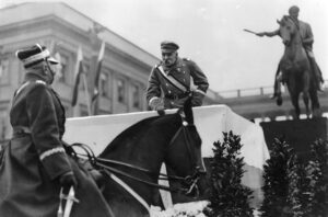 Józef Piłsudski pochyla się z podestu i zwraca do oficera na koniu. W tle pomnik konny Józefa Poniatowskiego.