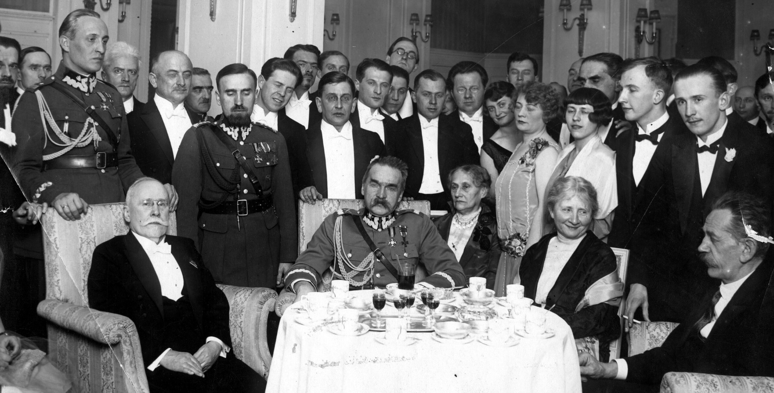 Przy stole od lewej Wacław Sieroszewski i Józef Piłsudski. Przy stole siedzi też Jędrzej Moraczewski. Za nimi stoi grupa odświętnie ubranych osób, wśród nich m.in. Andrzej Strug i 