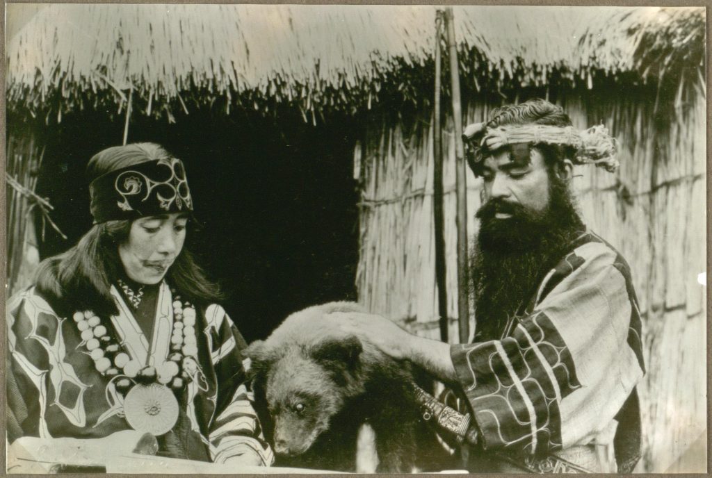 kobieta i mężczyzna w tradycyjnych strojach Ajnów przyglądają się niedźwiadkowi stojącemu przed nimi 