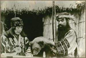kobieta i mężczyzna w tradycyjnych strojach Ajnów przyglądają się niedźwiadkowi stojącemu przed nimi